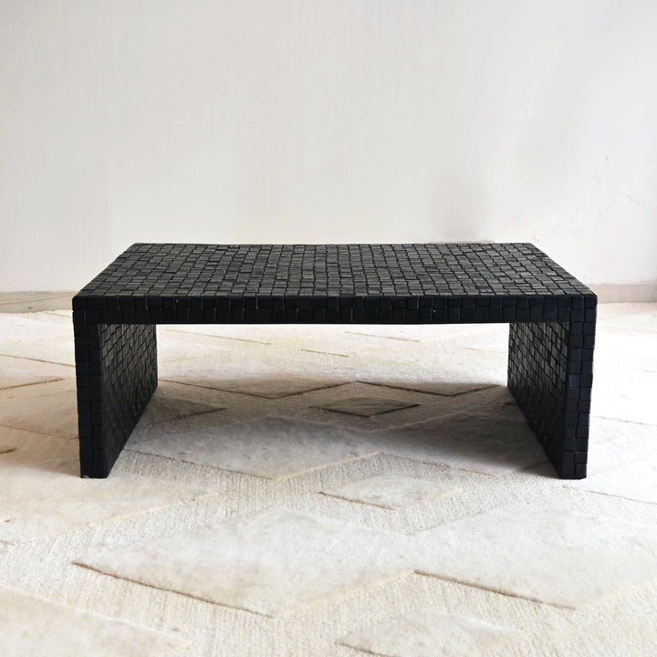 Black woven coffee table on a light wooden floor with a white wall background