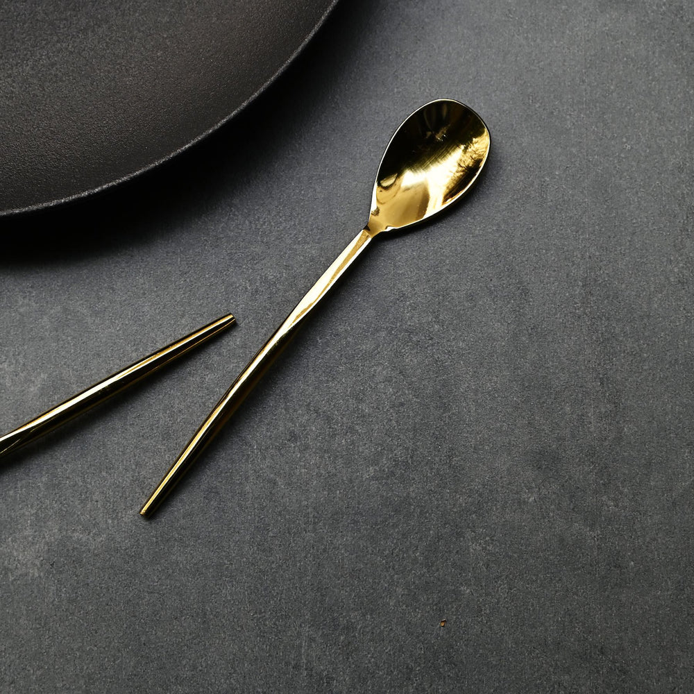 Aurum gold dessert spoon with a slender handle beside a dark plate.