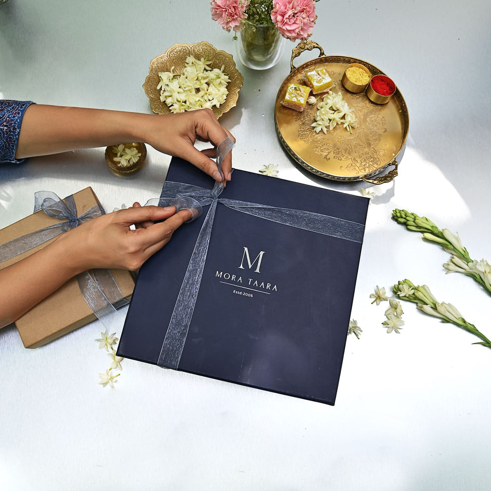 Navy blue gift box with 'Mora Taara' branding being tied with a ribbon on a white surface with decorative trays.