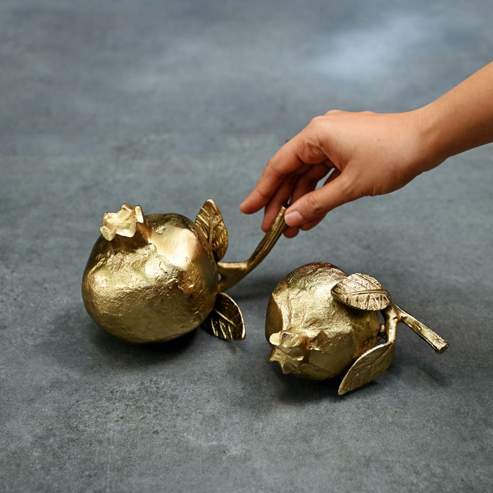 Gold decorative pomegranate sculptures on a gray surface with a hand interacting with one of them.