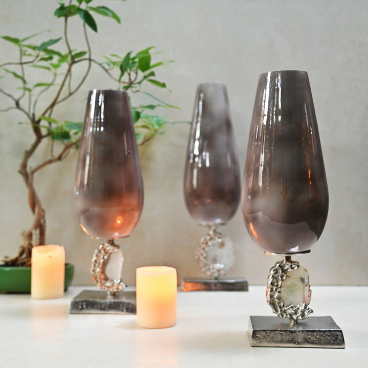 Three decorative glass vases with metallic bases on a surface with candles and a plant in the background.