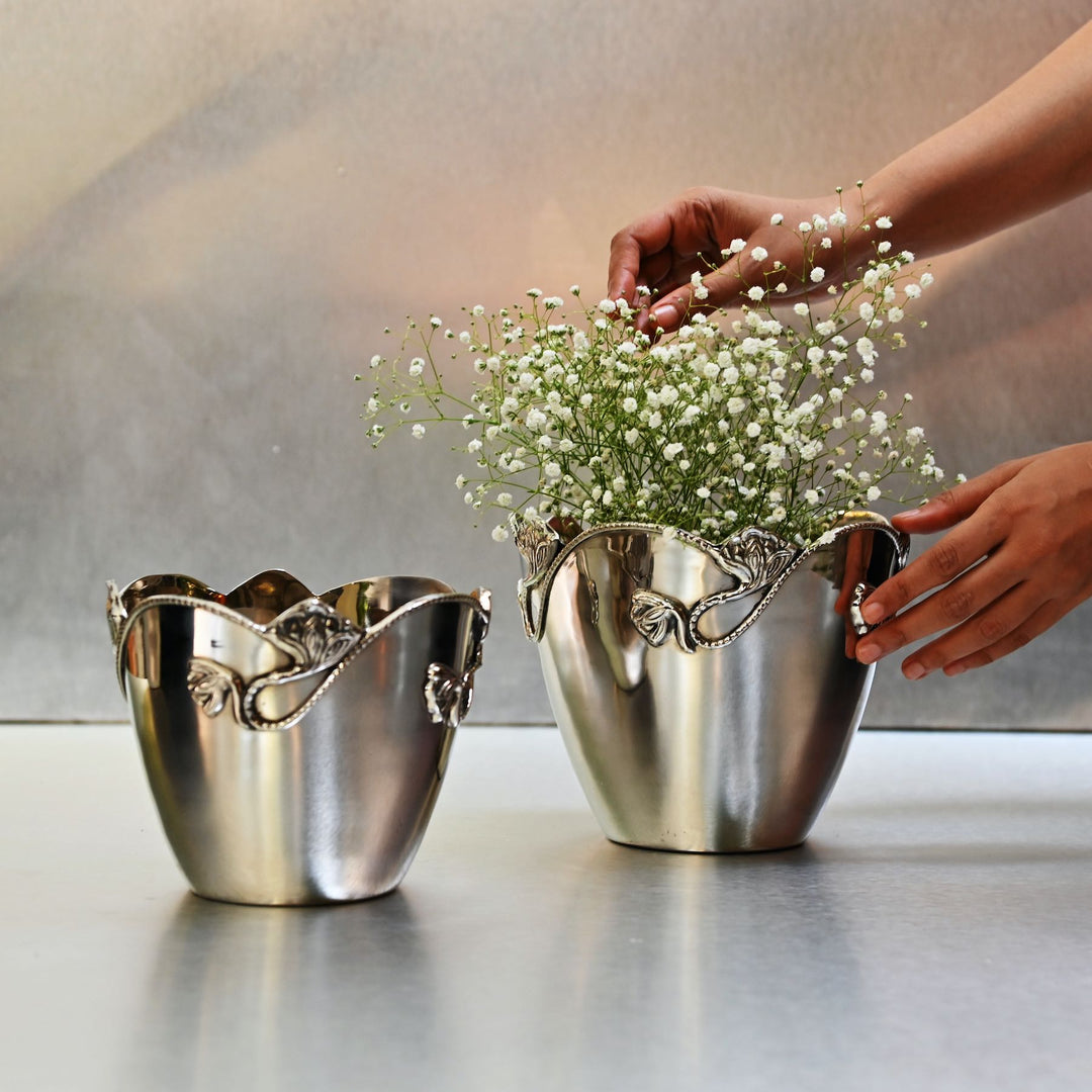Person arranging flowers in a decorative silver planter on a reflective surface.