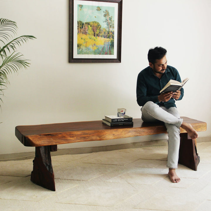 Man reading a book on a wooden bench in a room with a painting on the wall.
