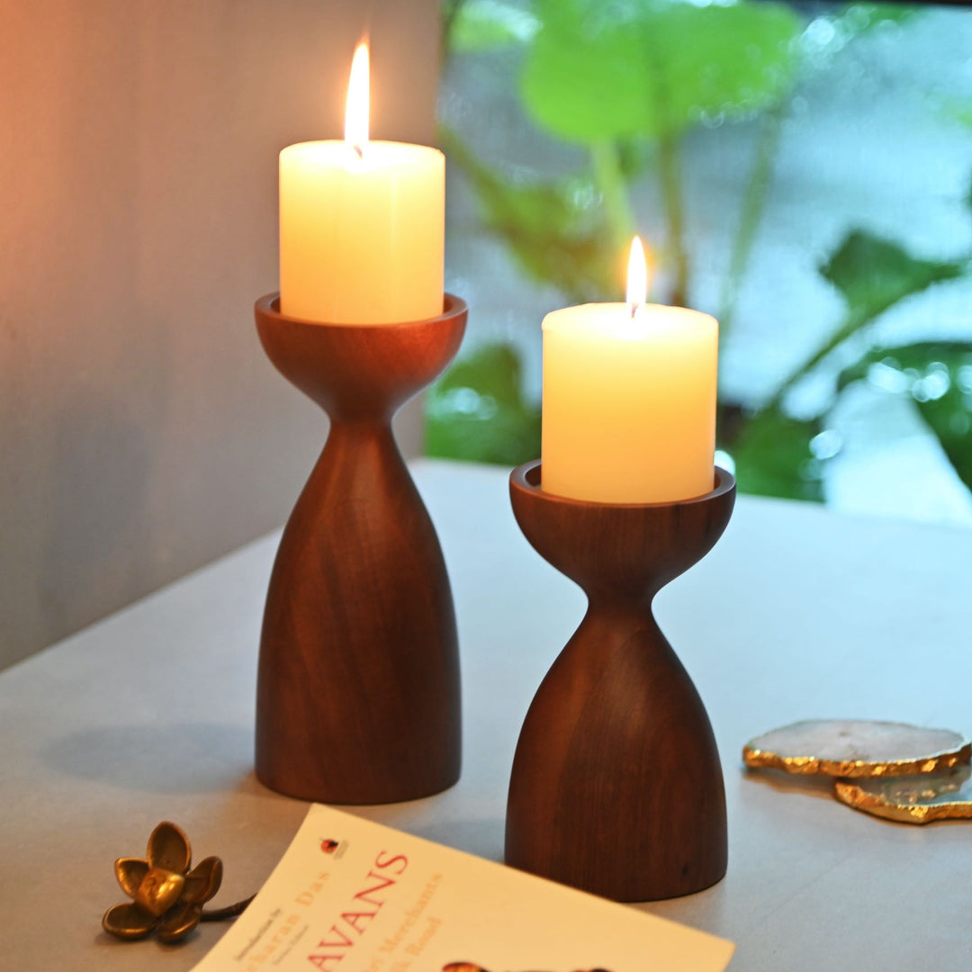 Two wooden candle holders with lit candles on a table with a blurred plant background
