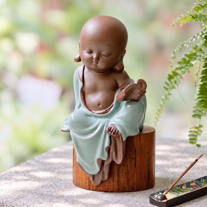 Celadon ceramic monk figurine holding a book, displayed on a wooden pedestal beside a fern.
