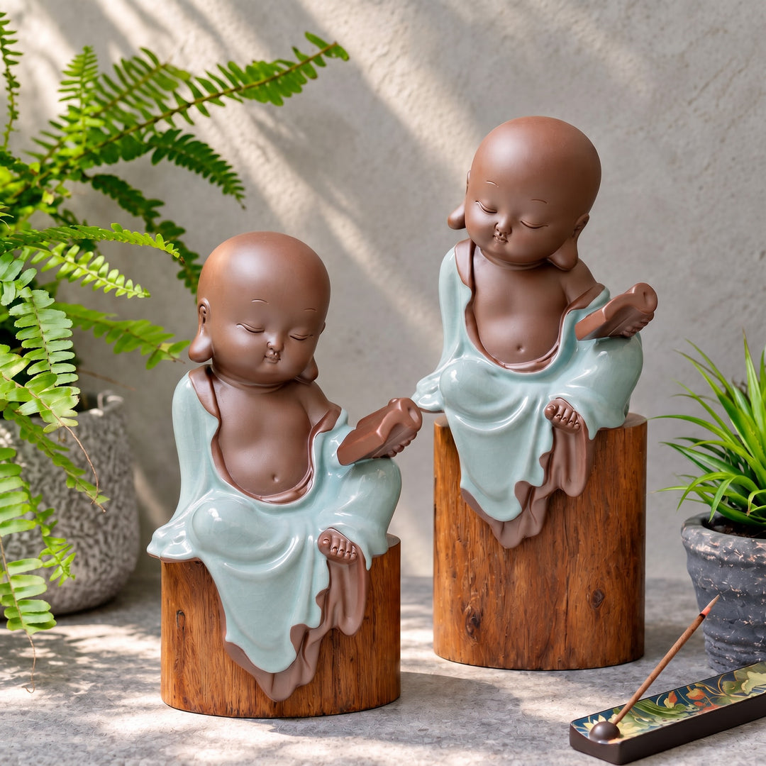 Two celadon ceramic monk figurines displayed on wooden pedestals among green plants.