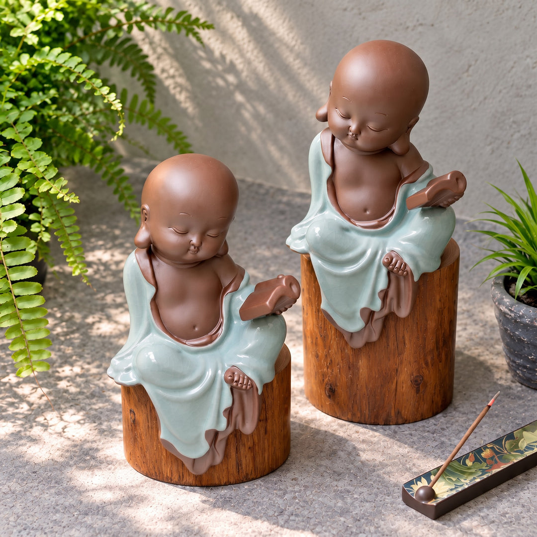 Angled view of two celadon monk figurines on wooden pedestals beside a fern and incense holder.