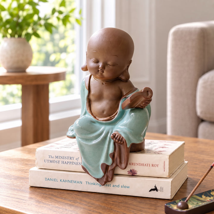 Celadon ceramic monk figurine on stacked books beside an armchair in a sunlit reading corner.