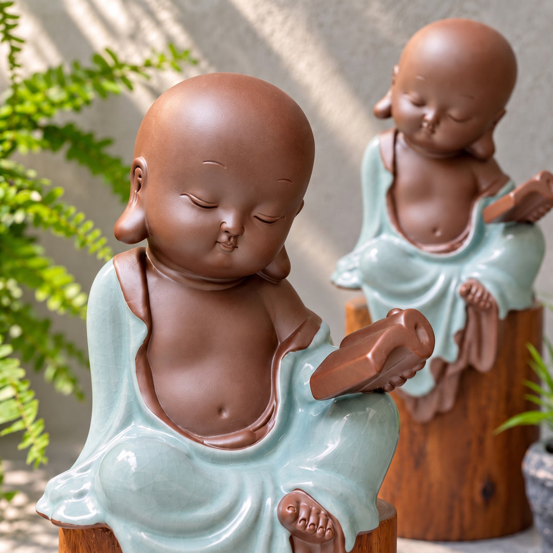 Close-up of a celadon monk figurine holding a book, with a second figurine softly blurred behind.