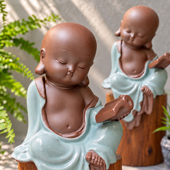 Close-up of a celadon monk figurine holding a book, with a second figurine softly blurred behind.
